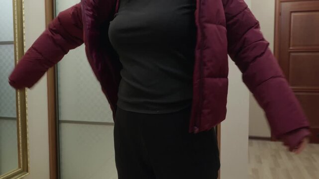 Young woman in dark outfit putting on padded jacket indoors near mirror, focused calm mood symbolizing preparation to go out and daily routine.