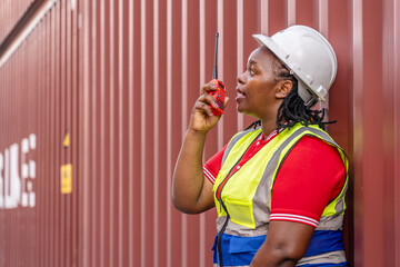 African American female logistics worker using a walkie-talkie. Industrial engineer in safety vest and hard hat supervising operations near a red shipping container terminal background.