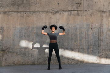 Determined athlete in boxing gloves flexing her biceps with confidence, casting a strong shadow on a concrete wall