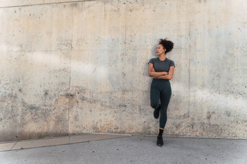 Confident sportswoman relaxing with crossed arms against concrete wall, looking away, after finishing her daily workout