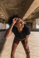 Exhausted female athlete resting under a bridge, touching her forehead and knee after a workout