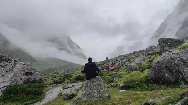 Peaceful moment of a man sitting amidst giant boulders and mist in the Vasudhara Falls trekking valley.