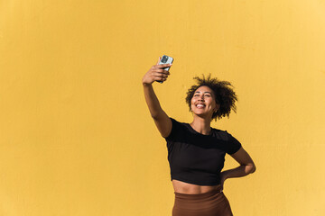 Smiling young woman taking a selfie with her smartphone against a vibrant yellow wall
