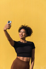Smiling woman taking a selfie with her smartphone in front of a vibrant yellow wall