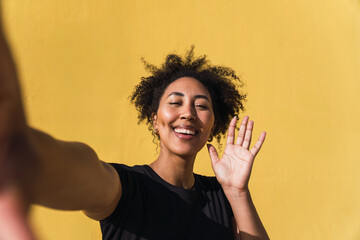 Cheerful young woman taking a selfie and waving her hand against a vibrant yellow background