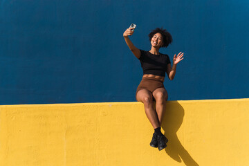 Sporty woman on a yellow wall video chatting on her smartphone with a blue background, flashing a smile