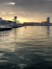 Sunset over Barcelona waterfront with boats, golden sky, and iconic modern architecture. Sunset at Barcelona Port Vell with Rambla de Mar bridge and W Hotel under a dramatic cloudy sky.