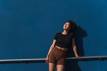 Fit woman in sportswear enjoying the sun after exercising outdoors, leaning against a blue wall