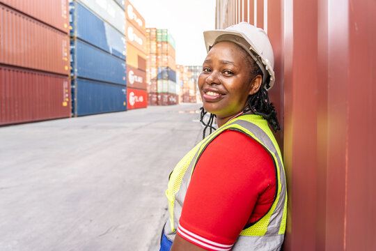 Happy African American female worker standing in a shipping yard. Portrait of a logistics engineer wearing a safety vest and hard hat leaning against a cargo container at a busy commercial port. - Powered by Adobe