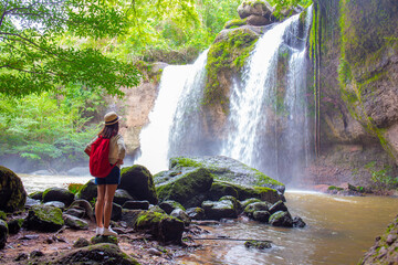 Female tourist wearing a hat and carrying an adventure backpack stands in front of Heo Suwat Waterfall, Khao Yai National Park, Nakhon Ratchasima, Thailand.