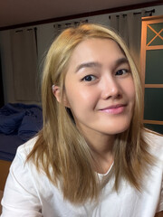 Asian Thai woman with blonde hair, smiling and taking selfie while sitting in cozy and warm bedroom.