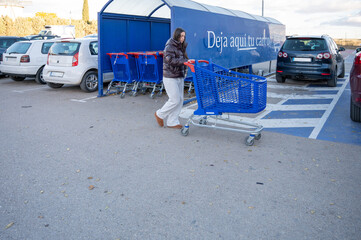 Teenage girl returning empty cart to designated area in supermarket parking lot on a cold sunny day, wearing winter jacket and casual sweatpants after grocery shopping