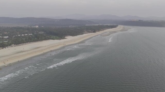 Evening aerial view of Mobor Beach, South Goa, showing white sand coastline and gentle ocean waves bordered by lush coastal vegetation with coconut palms and distant hills under soft light.