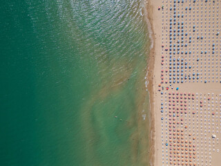Top-down aerial view of a sandy beach with turquoise sea and geometric rows of sun umbrellas and loungers. Minimal summer composition showing contrast between nature and organized leisure