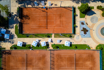 Top-down aerial view of clay tennis courts separated by a walkway with umbrellas and seating. Clean geometric layout, symmetry, warm colors, outdoor sports and recreation concept