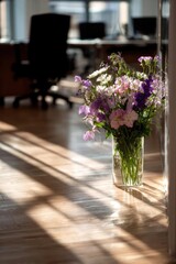 Fototapeta premium A glass vase of pink, purple and white flowers sits on a sunlit wooden floor in an office, with blurred desks in the background. Concept Office flowers, Glass vase, Sunlit wood floor