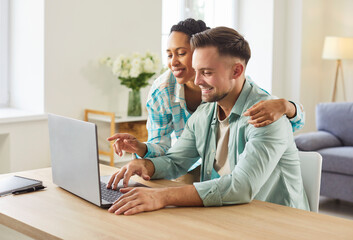 Happy couple sit at home, using a laptop together to plan finances while smiling at the screen and discussing options in a cozy living room. They are planning finances online together today.
