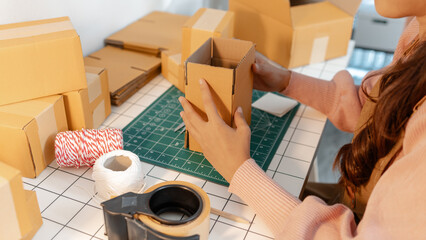 A young woman is carefully assembling cardboard boxes at a neatly organized workspace. The desk is filled with packing materials, tape, and string, reflecting focus and small business activity.