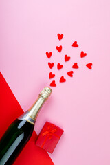 Champagne bottle splashing red heart shapes by gift box on pink background