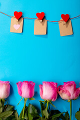 Red heart shapes on clothespins hanging from clothesline over pink roses against blue background