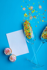 Champagne flutes with confetti by cupcakes and blank paper with copy space on blue background