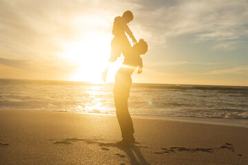 Naklejka premium Side view of back lit father holding aloft son on shore at beach against sky during sunset