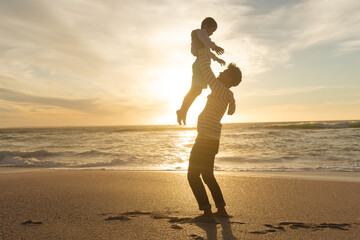 Naklejka premium Side view of playful father holding son aloft on shore at beach against sky during sunset