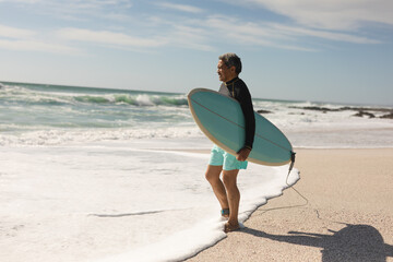 Retired senior man carrying surfboard walking on shore at beach during sunny day