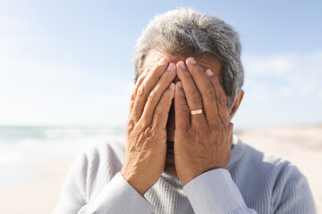 Close-up of worried senior man covering face with hands at beach against sky on sunny day