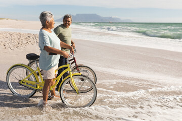 Obraz premium Full length of senior multiracial couple with bicycles standing on shore at beach during sunny day