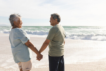 Happy retired multiracial senior couple holding hands while looking at each other on sunny beach