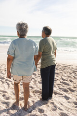 Obraz premium Full length rear view of multiracial senior couple standing on sand at beach during sunny day