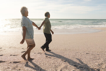 Obraz premium Full length side view of multiracial senior couple holding hands while walking on shore at beach