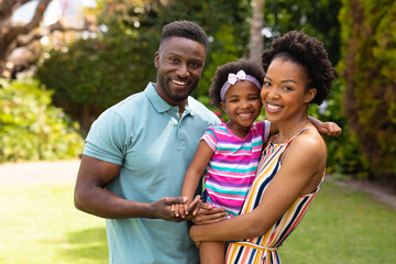Portrait of smiling african american parents carrying cute daughter standing at garden
