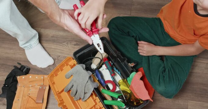 Boy sitting on floor hands pliers from toolbox to father during home repair task. Skillful child assists father by selecting correct tool for furniture work