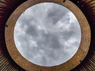 Circular ceiling opening reveals cloudy sky framed by corrugated metal and OSB structure with perimeter fixtures.