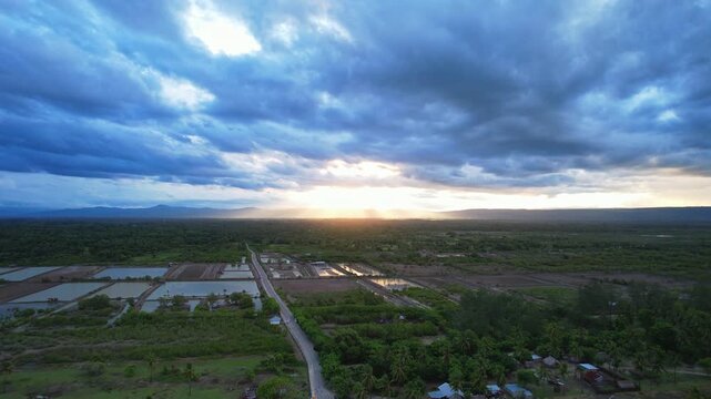 Aerial drone footage of the main aquaculture area, forest, in Malaka regency, Timor island, Indonesia, with vegetable fields in a flat valley, hills behind with a strong sunset, in a dark cloudy time