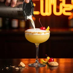 Fototapete Bar shaker pours a frothy yellow almond cocktail into a coupe glass rimmed with crushed amaretti cookies. Background: a dark vintage bar with warm amber neon. A bright red cherry and lemon twist provide c  © ❤  Nancyvoz68❤ 