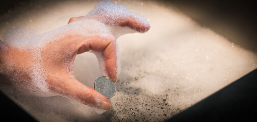 Hand Holding Coin in Pinched Fingers Gesture Over Foamy Sink &mdash; Money Laundering Concept