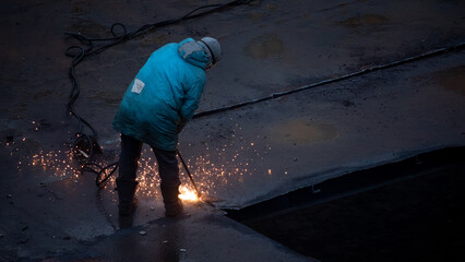 Single industrial worker cutting metal roof panels with power tool, producing bright sparks during construction or repair work at site