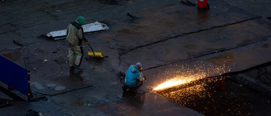 Two industrial workers cutting metal roof panels at construction site, bright sparks flying during repair or demolition work in wet conditions.