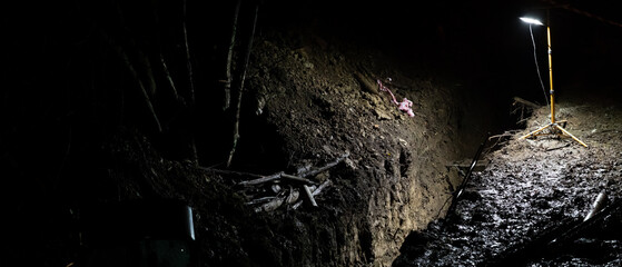 Dark outdoor excavation site at night with muddy trench, exposed soil layers and portable work lamp illuminating construction or utility groundwork.