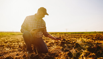 An experienced farmer with a clipboard checks the soil quality before sowing. An agronomist in a checkered shirt works in a field, holding a sample of black soil. Concept of gardening and ecology.