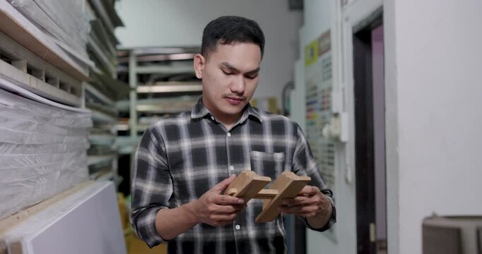 Asian adult male woodworking business owner compares multiple wood samples, rotates pieces, judges hardness grain in small workshop, guiding artisan wood selection process for interior furniture work