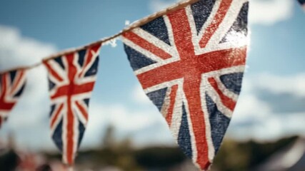 A vibrant string of festive bunting, adorned with a classic red, white, and blue cross and diagonal stripe pattern, gently sways against a bright blue sky. The triangular fabric flags, with their text