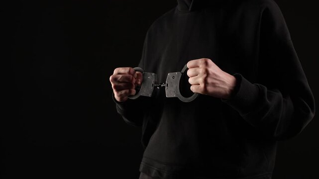 A person in a dark hoodie holds a pair of open handcuffs tightly in both hands, framed against a black background, suggesting restraint or confrontation