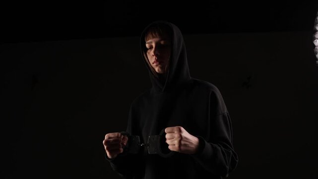 A person in a black hoodie stands in dim lighting, holding open handcuffs with a serious expression, evoking a mood of conflict or reflection