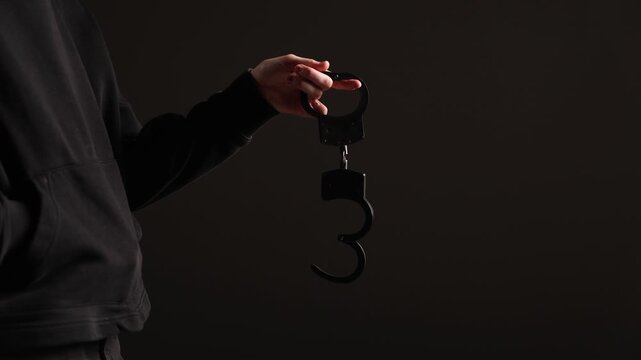 A person in a dark hoodie holds an open pair of black handcuffs against a dark background with ample copy space, creating a mysterious and dramatic atmosphere