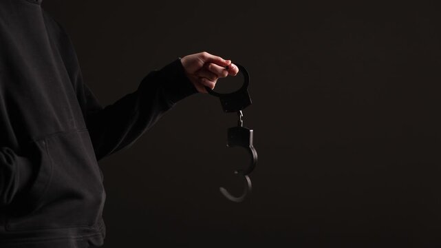 A person in a dark hoodie holds an open pair of black handcuffs against a dark background with ample copy space, creating a mysterious and dramatic atmosphere
