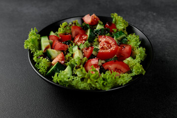 Vegetable salad of tomatoes, cucumbers, onions, lettuce dressed with aromatic dressing in a black plate on a gray background